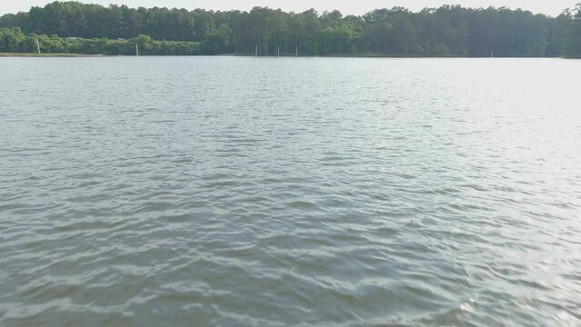 aerial footage of the rippling waters of Lake Acworth surrounded by lush green trees, grass and plants with cars driving on the road and blue sky with clouds at Proctor Landing Park in Acworth Georgia