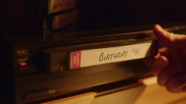 Close Up of a Person Inserting a VHS Cassette in a Player with Nostalgic Birthday Footage from Home Video Camera. Retro Nineties Technology Concept. Old VCR Shot with Shallow Depth of Field and Bokeh.