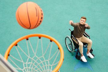 Concentrated young man with paralyzed legs sitting in wheelchair and biting lip while throwing ball...