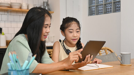 Asian mother helping her daughter doing homework, learning online at virtual class on laptop computer at kitchen table