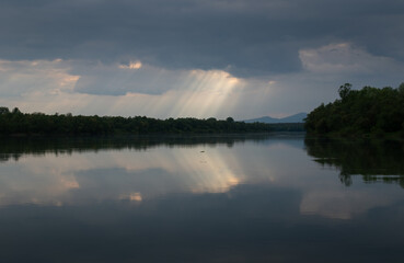 Sun ray piercing through clouds over river, forest on river bank and mountain silhouette in distance