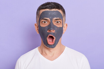 Horizontal shot of shocked astonished man with clay mask on his face, receiving spa treatments,...