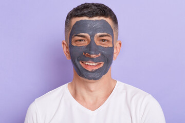 Indoor shot of happy smiling man with clay mask on his face, receiving spa treatments, doing...