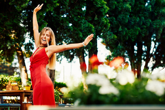 Happy Young Woman In A Red Dress With Her Arms Raised In The Summer Garden