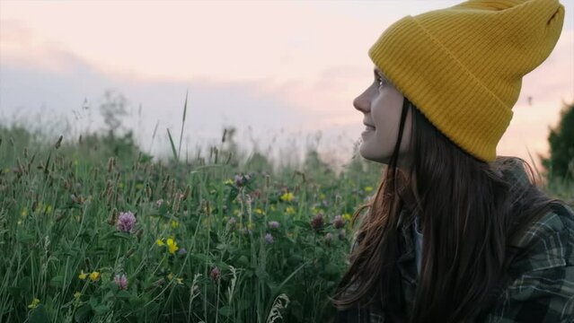 Happy young woman with long hair, checkered shirt and knit yellow beanie sitting in grass on background colorful sky during sunset, embrace nature elements, travel blogger lifestyle. Slow-motion