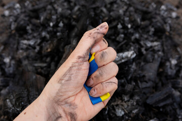 the symbol of ukraine is clutched in the hand against the background of coals from the conflagration. ukraine on fire, struggle for freedom in ukraine. yellow blue ribbon of the flag of ukraine