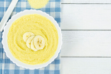 Corn porridge for a baby from ground cereals in a bowl with a banana on a blue napkin on a white...
