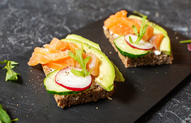 Two open sanwiches with dark rye bread, avocado, smoked salmon and radish, black background