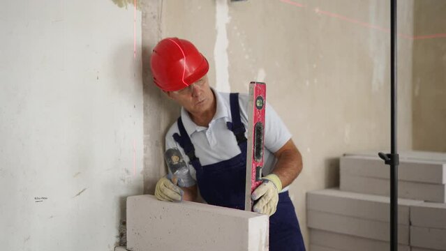 Bricklayer using rubber mallet hammer to tap and level concrete blocks wall. Contractor uses precise spirit bubble and laser level for brickwork. Worker constructs a wall in new apartment real estate.