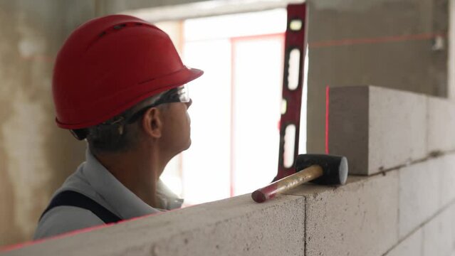 Bricklayer using rubber mallet hammer to tap and level concrete blocks wall. Contractor uses precise spirit bubble and laser level for brickwork. Worker constructs a wall in new apartment real estate.