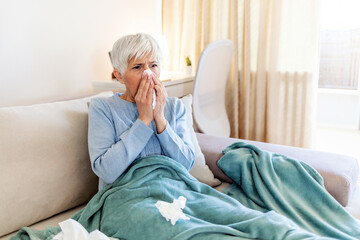 Shot of a senior woman blowing her nose with a tissue at home. Senior woman blowing her nose while feeling sick at home.