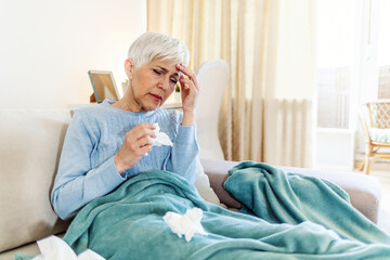 Sick mature woman sitting on sofa with headache under the blanket. Senior woman caught cold or flu...