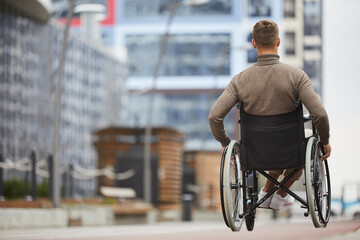 Fototapeta na wymiar Rear view of young man in brown turtleneck pushing hand rims of wheelchair while riding wheelchair in city