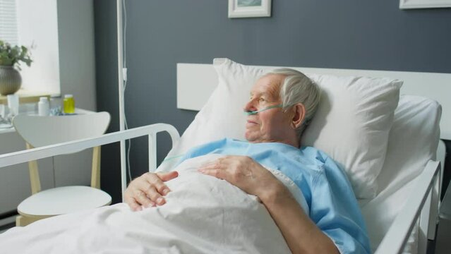 Zoom in shot of sick elderly man with nasal cannula lying on bed in hospital ward and looking through window