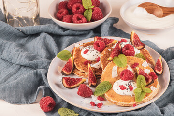 Pancakes with raspberries, figs, yogurt, coconut zest, honey and mint leaves on a plate.
