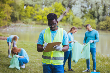 Medium shot of young African volunteer with clipboard. He is with his team outside in the park, with bags, gloves and buckets, cleaning and collecting garbage