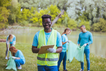 Medium shot of young African volunteer with clipboard. He is with his team outside in the park, with bags, gloves and buckets, cleaning and collecting garbage