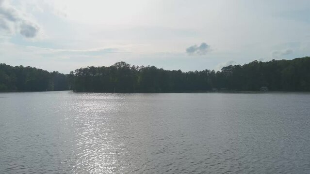 aerial footage of the rippling waters of Lake Acworth surrounded by lush green trees, grass and plants with cars driving on the road and blue sky with clouds at Proctor Landing Park in Acworth Georgia