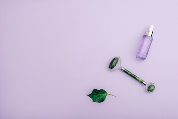 Cosmetic serum, jade facial roller and green leaf on purple background. Skincare, spa and wellness concept. Top view, flat lay, copy space