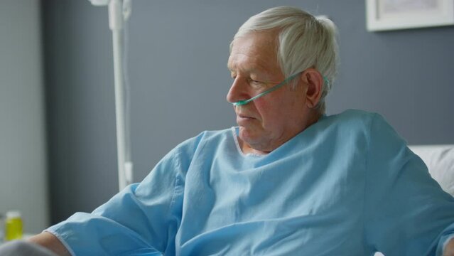 Tilt up shot of elderly male patient with nasal cannula getting up, taking off blanket and sitting on hospital bed while feeling better during recovery