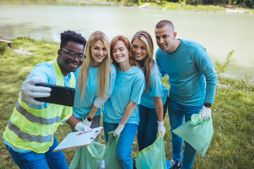 A multiethnic group of volunteers is resting after cleaning the city park. They take selfies and laugh. Happy to have done the job.