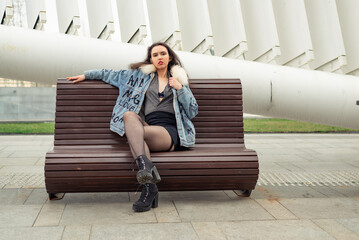Obraz premium A beautiful girl in a denim jacket and a black short skirt sits on a bench in the city