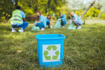 A recycling bin stands in a meadow full of plastic bottles, while in the background is a group of...