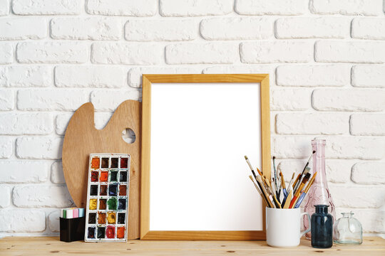 Photo frame as easel with artist's tools on wooden table against white brick wall