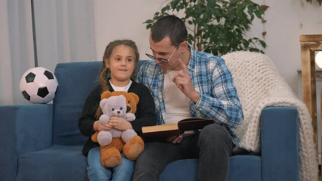 Happy family. A girl with toys listens to dad story. Father reads book to his daughter. Go to sleep. Family training in quarantine. Homeschooled girl. Dad reads a book to his daughter before bedtime