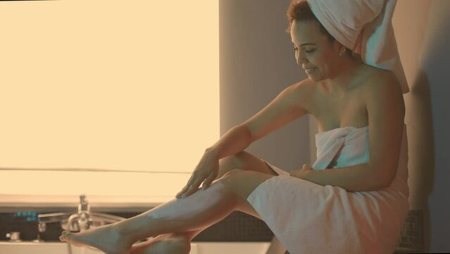 woman sitting on the edge of the bathtub spreads cream on her legs - warm light