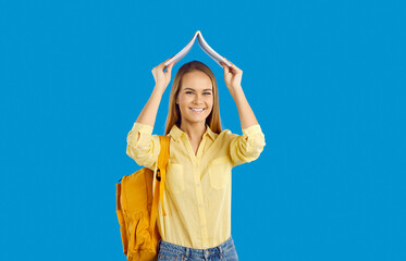 Portrait of smiling Caucasian teenage girl with backpack hold book over head excited about studying. Happy millennial female student with textbook isolated on blue studio background. Education.