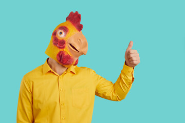 Funny man with chicken head raises his thumb up demonstrates you OK gesture, consent or his support. Eccentric man in yellow shirt and with rubber mask of chicken on his head on light blue background. - obrazy, fototapety, plakaty