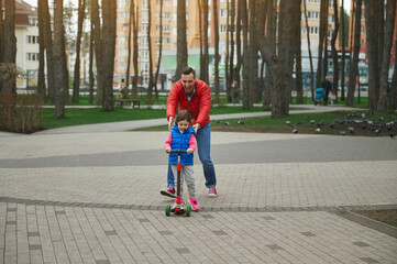 Cheerful dad running beside his cute daughter riding a push scooter, enjoying day off in a city park. Father's Day, happy childhood concept