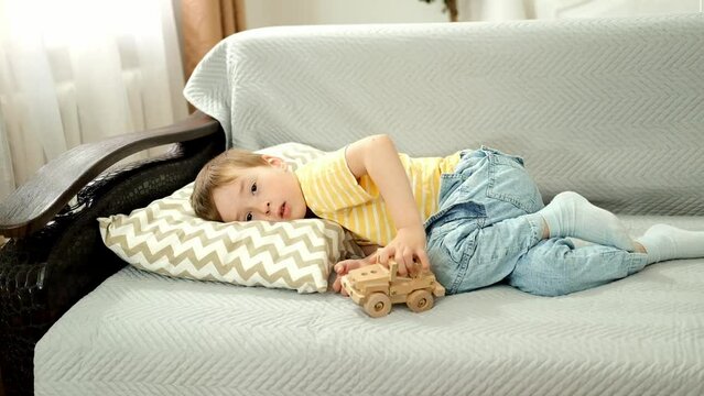 Sick ill Caucasian child is lying in bed and playing with toy car. Boy is lying on sofa, is bored, watching TV.