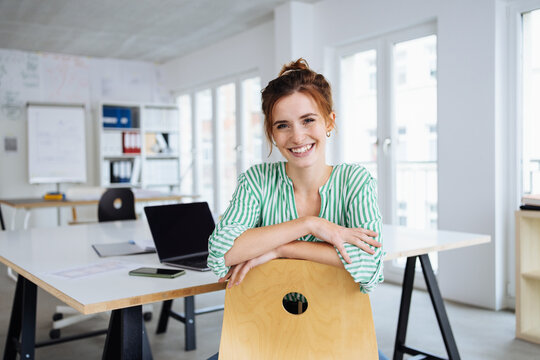 Confident young businesswoman looks at camera laughing and leans on her desk chair
