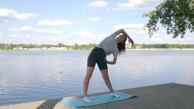 a young girl is doing yoga on the shore on the yoga mat of the river in the evening