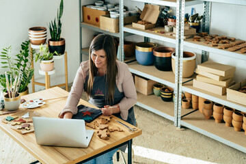 From above photo of happy woman artisan manufacturing handmade ceramics Christmas decoration while sitting at table in her studio and watching online video tutorial on a laptop