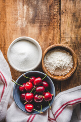 vista cenital de cuencos con cerezas, harina y azúcar sobre una mesa de madera rústica, para preparar un postre