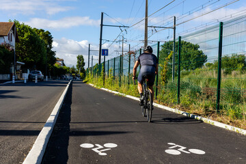 Piste cyclable avec un cycliste le long d'une route, la piste est séparée par un trottoir, le sol possède un marquage blanc pour indiquer le sens des vélos - obrazy, fototapety, plakaty
