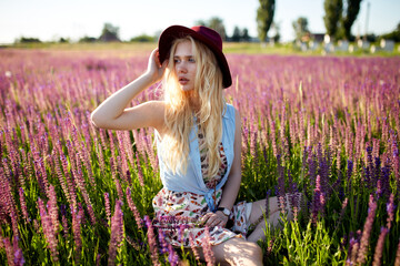 Profile portrait of a young girl with shiny blond hair is sitting on a field of flowers, wearing a hat.