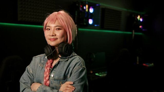 Cute young Asian girl in pink wig posing to a photo in headset holding black game controller and smiling. High quality 4k footage