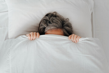 Top view tired caucasian woman with gray hair is covered with blanket lying on bed in bedroom. Concept depression, laziness, frustration, female mental health