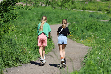 Two kid girls on electric scooters go downhill in summer park