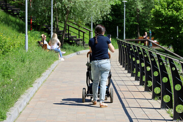 Woman with baby pram walking with smartphone on sidewalk. Mother with child, leisure in summer park