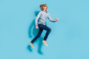 Fototapeta na wymiar Full length photo of shocked excited young guy dressed jeans shirt eyewear jumping high running fast isolated blue color background