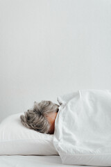 Concept depression, frustration, laziness, stress. Side view of tired woman with gray hair covered with blanket lying on bed in bedroom. Vertical view, copy space
