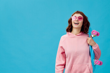 Funny joyful pretty redhead lady in pink hoodie sunglasses with penny board smiling posing isolated on blue studio background. Copy space Banner Offer. Fashion Cinema. Holiday activity