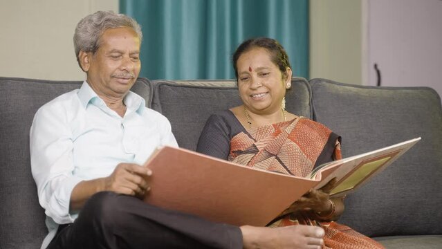 Handheld shot of happy senior couple laughing by looking into to marriage photo album - concept of old memories, nostalgic and family bonding