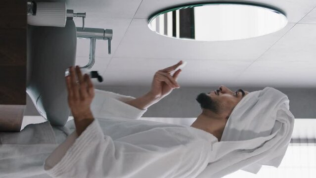 Handsome arabic idian bearded man in bath robe with towel on head uses liquid skin care cosmetic cleans face with tonic-water looking in mirror rubbing cheeks with cotton pad with anti acne product