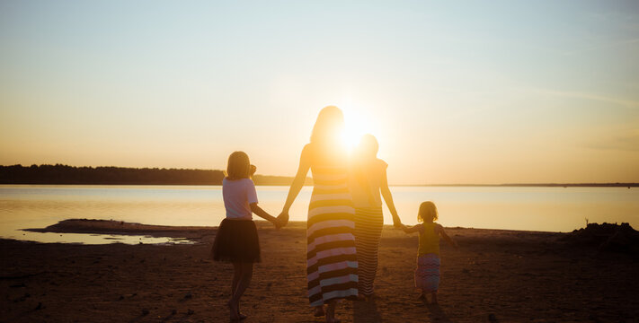 Silhouettes Of Children And Mother Walking Hand In Hand On The Sea Beach To Sunset Light. Good Mood And Younger And Older Generation Pastime. Beautiful Landscape. Family Love. Woman Friendship. Peace